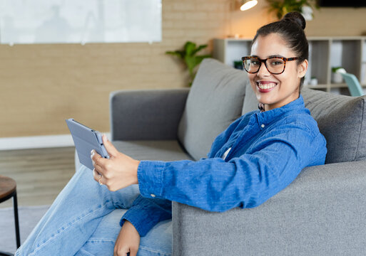 Woman sitting on sofa holding tablet sporting glasses in living room with potted plant, copy space