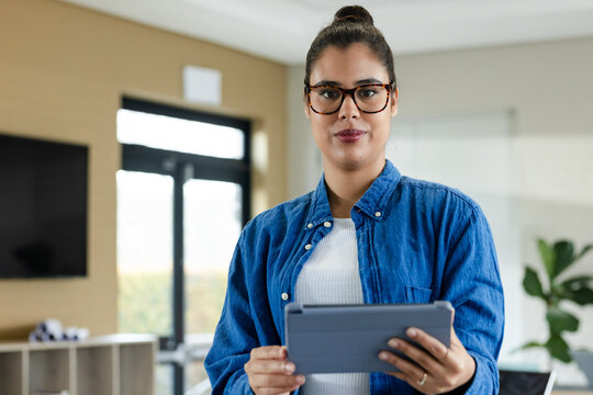 Hispanic woman holding tablet while wearing tortoiseshell eyeglasses in modern office with TV