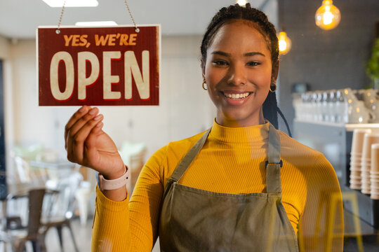 African American woman barista holding hanging YES WE'RE OPEN sign behind coffee shop glass window