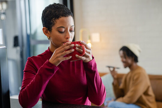 Diverse friends sitting at kitchen counter holding red mug and smartphone by sofa lamp, copy space