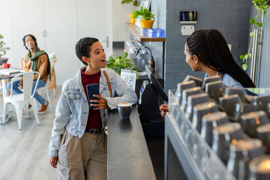 Female customer placing order using phone at counter in cafe with barista operating POS terminal