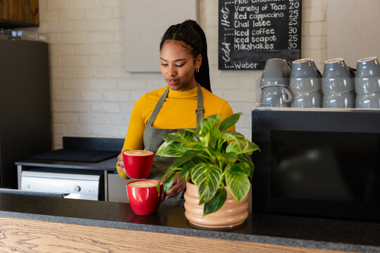 Adult African American woman barista placing two cups on wooden counter at cafe by chalkboard menu