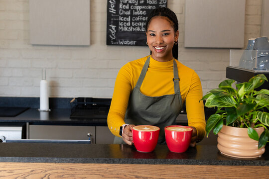 African American female barista smiling and presenting two red latte mugs at coffee shop counter