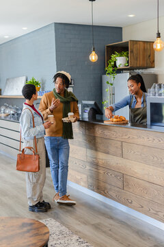 Diverse couple ordering paper cups, barista serving pastries at cafe counter hanging Edison bulbs