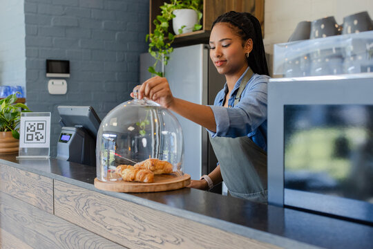 African American woman in apron lifting glass dome on cafe counter showing croissants by POS system