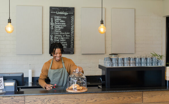 African American male barista lifting glass cover and serving croissants behind coffee shop counter