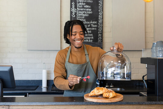 Smiling African American male barista lifting glass cloche dome over croissants at cafe counter