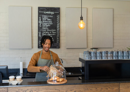 African American male barista lifting glass dome over croissants behind counter by takeaway cups