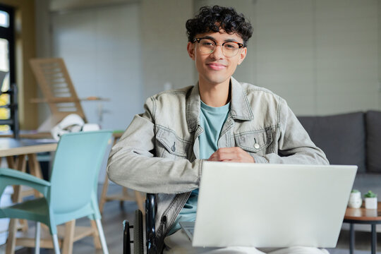 Man sitting in wheelchair using laptop in modern lounge wearing denim jacket and eyeglasses