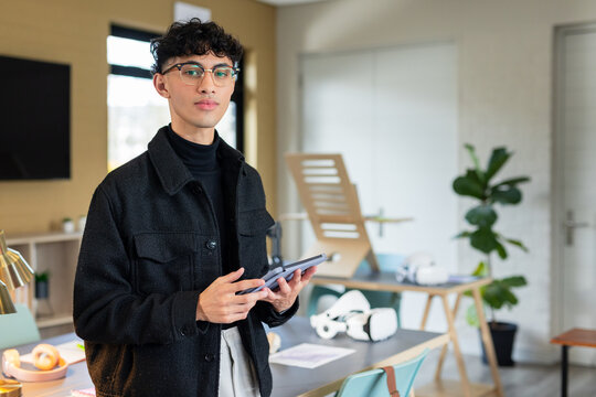 Asian man standing holding tablet in modern office surrounded by VR headsets and prototypes