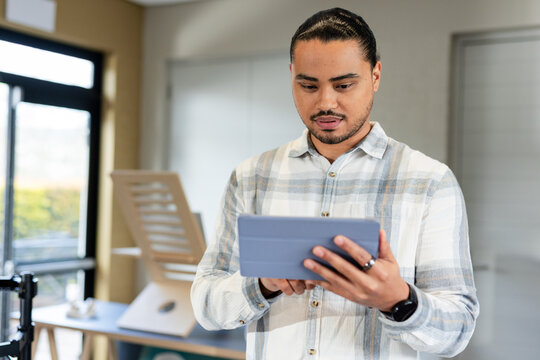 Asian man standing in design studio holding tablet with smartwatch, examining architectural models
