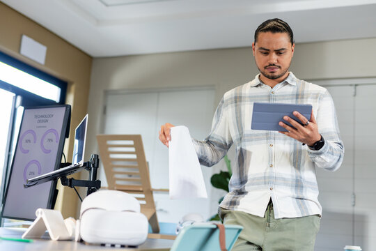 Asian American man holding printed graphs and reviewing tablet beside monitor at office desk