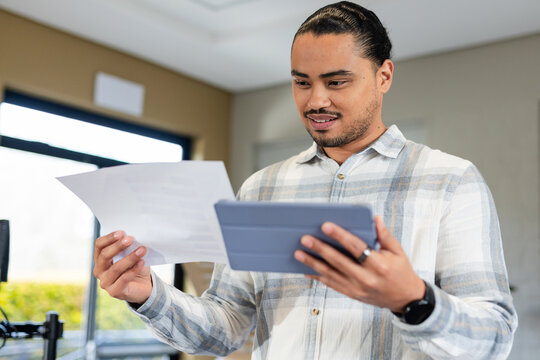 Asian man wearing plaid shirt and smartwatch using tablet and comparing paper in office, copy space