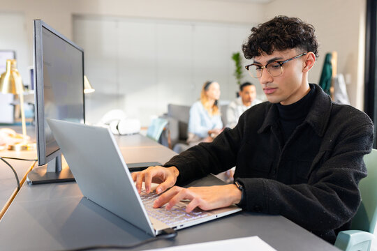Man in early 20s typing on silver laptop at co-working office desk with large external monitor