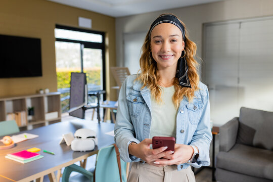 Woman standing in office holding smartphone smiling by table with VR headset and notebooks