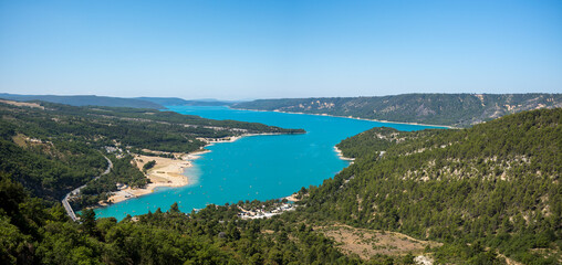 Fototapeta premium Panoramic view of Lac de Sainte-Croix showing vivid turquoise water bordered by sandy shores and dense green hills beneath a bright blue sky.