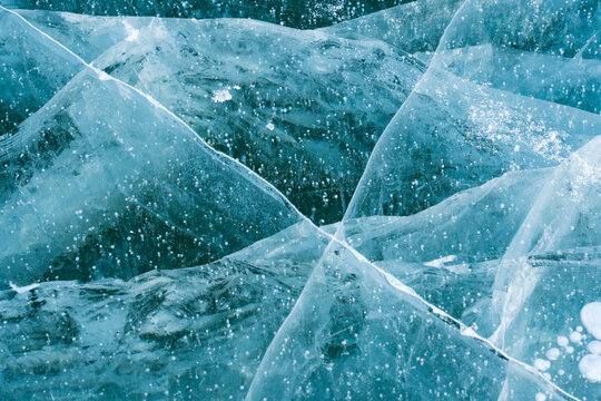 Abstract top-down view of deep blue frozen lake ice featuring intricate white cracks, fractures, and frozen bubbles.