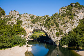 Fototapeta premium A striking natural limestone arch rises above a calm river, surrounded by rugged cliffs and dense green trees in the Pont d'Arc area of Ardeche under a clear blue sky.