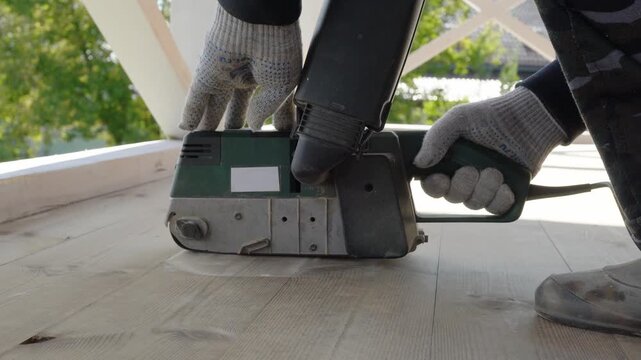 Carpenter smooths wooden floor with electric sander outdoors on sunny day