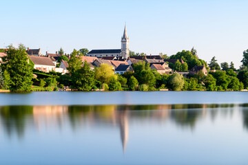 Fototapeta premium A tranquil pond reflects the church spire and traditional houses of Dampierre en Burly, surrounded by lush greenery and bathed in warm summer sunlight. The smooth water surface and clear sky create a