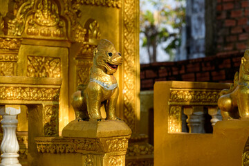 Fototapeta premium Close-up of a gold-painted guardian lion sculpture with intricate detailing, set against ornate golden temple architecture at Phnom Sampov in Battambang province, Cambodia. The scene is illuminated by