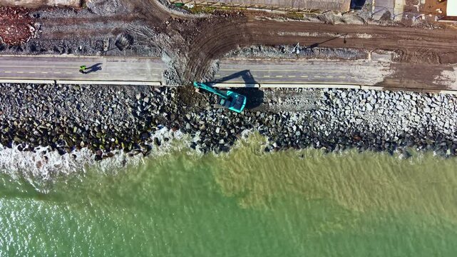 Workers use heavy machinery to move rocks and materials along a road by the water. The construction area shows a mixture of dirt, rocks, and water at nearby shoreline.