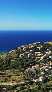Scenic aerial shot of Corbara village perched on a hillside overlooking the deep blue Mediterranean Sea under a clear sky. Lush greenery and traditional buildings create a picturesque coastal