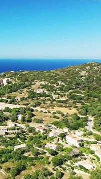 Scenic aerial shot of Corbara village nestled in lush hills with the Mediterranean Sea in the background, under a clear blue sky.