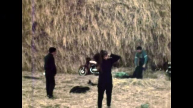 Biker friends finishing rest stop near haystacks in countryside. Riders prepare to continue motorcycle journey after rural break. Road trip in summer. Old film. Archival retro 1970s. Vintage archive