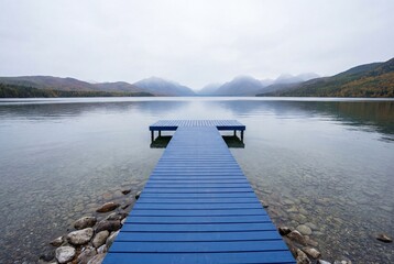 Long blue wooden dock on tranquil lake water with foggy mountains and pebble shore in background