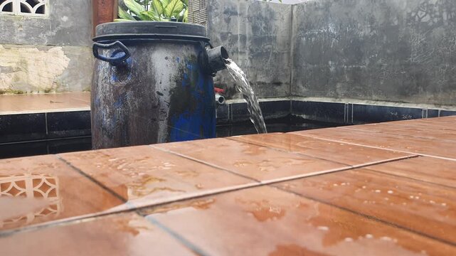 Blue plastic drum used as a pond filtration system with water flowing into a dark tiled fish pond in garden