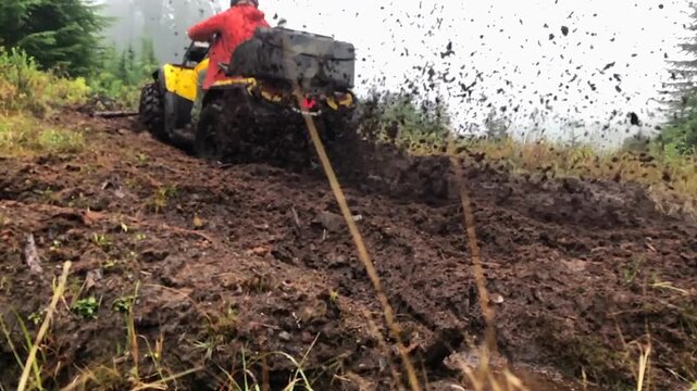 Man riding a yellow ATV trying to climb up a steep dirt hill, unsuccessfully, and spraying dirt and debris everywhere towards the camera.