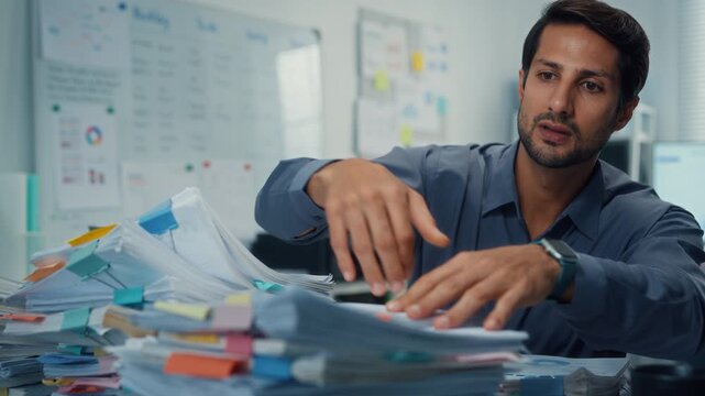 Overworked young Latin businessman reading document in large stack of paperwork files at office desk. Tax audit deadline, accounting stress, corporate burnout, bookkeeping, bureaucracy concept.