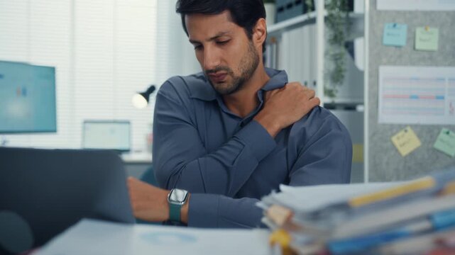 Young Latino businessman suffering shoulder pain and neck ache at office desk with computer. Office syndrome, muscle massage, overworked stress, bad posture, and health insurance injury concept.