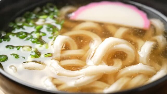 Close-up of delicious hot udon noodles submerged in dashi broth, garnished with chopped green onions and traditional pink-rimmed kamaboko fish cake in a Japanese restaurant setting.