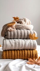 Stacked warm sweaters in autumn colors with scattered fallen leaves on white textile
