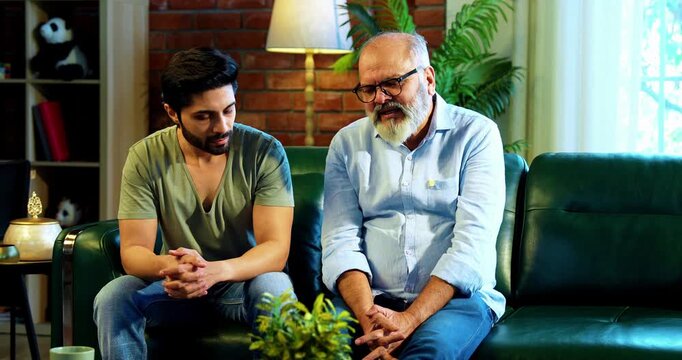 Indian young son and senior father discussing serious life topic while sitting on sofa in modern living room at home, sharing personal experiences, wisdom emotional bonding between two generations