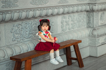 Adorable little Asian girl in traditional Chinese dress holding red envelope angpao, sitting on wooden bench at shrine, celebrating Chinese New Year festival, Lunar new year