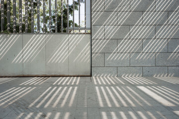 Fototapeta premium Sunlight filters through a metal fence casting diagonal shadows on stone walls and concrete ground