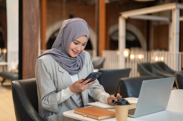 Asian muslim woman student looking at phone writing in book aside laptop and coffee sitting in cafe.