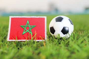 Morocco national flag beside a classic soccer ball on green stadium grass in daylight. Concept of global tournament ambition, African football rise and national pride. © The Dell Shot by SJ