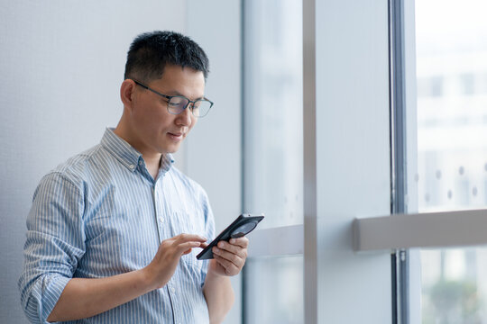 A middle-aged Asian man wearing glasses focuses intently on operating a smartphone by the window in a modern office setting
