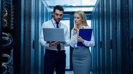 Tech professionals inspecting data server: Two dedicated tech professionals, immersed in a server room, meticulously examine a laptop and documentation amidst an intricate network of data servers.