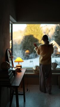 Woman practicing a calm morning self care ritual by the window in a cozy modern mountain house during winter. Soft natural light, snowy landscape outside and peaceful slow living atmosphere