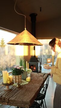 Woman enters the frame, drinks coffee and sits at a wooden table while a fireplace burns in the background. Warm light, cozy interior and calm atmosphere in a modern mountain house. Slow living and