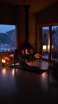 Woman practicing gentle yoga near a fireplace in a cozy mountain house during winter. Warm candlelight, calm atmosphere, slow living lifestyle, evening relaxation indoors