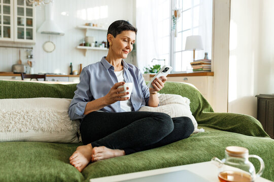 Relaxed pretty woman of 60s enjoying leisure time at home, sitting on green couch barefoot and in casual clothes drinking tea or coffee and using smartphone, chatting with chat bot