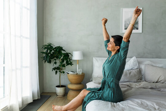 Side view of awake mature woman with closed eyes in blue soft cotton bathrobe stretching her hands and back sitting on bed facing panoramic window with curtains, feeling happy and well-rested