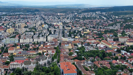 Obraz premium An aerial, wide-angle shot of Deva, Romania, showing the dense urban landscape with blocks of apartment buildings, older structures with red-tiled roofs and a main boulevard, set against rolling hills