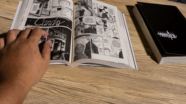 South Kalimantan, Indonesia - 02 16 2026 : Hand turning pages of Monster manga by Naoki Urasawa on wooden desk, interior comic panels visible under natural light creating casual reading